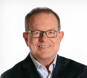 A middle-aged man with short hair, glasses, and a dark suit jacket smiles at the camera against a plain white background.